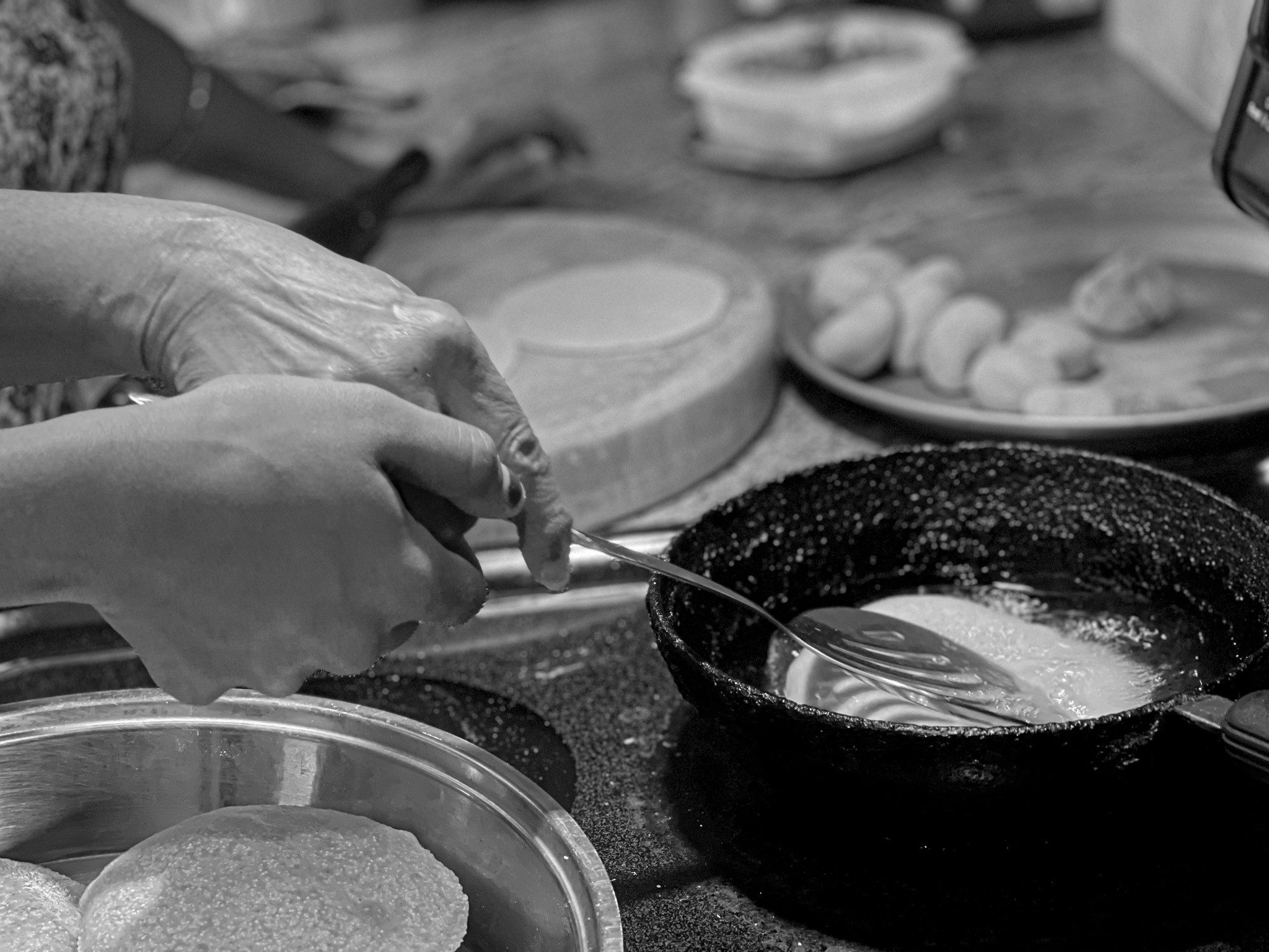 Two hands sharing a utensil above a frying pan, with dough and a rolling board behind them.